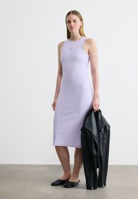 Γυναίκα στέκεται φορώντας μίντι φόρεμα χωρίς μανίκια σε ανοιχτό μωβ χρώμα, κρατώντας ένα μαύρο δερμάτινο μπουφάν στο δεξί της χέρι, φοράει μαύρα παπούτσια χωρίς κορδόνια.