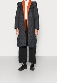 Person trägt einen schwarzen gesteppte langen Mantel mit Kapuze, einen orangefarbenen Strickschal, ein weißes Oberteil, schwarze weit geschnittene Hosen und schwarze klobige Stiefel.