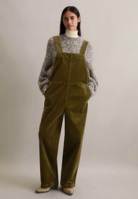 Vrouw in olijfgroene corduroy overall over een chunky gebreide trui en beige coltrui, staand met handen in de zakken.