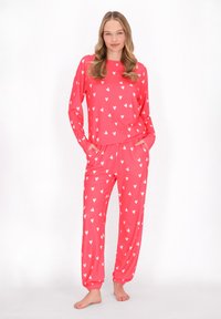 Roze pyjama set met lange mouwen en broeken met een overal wit hartpatroon. De stof is glad, met elastische manchetjes bij de enkels.