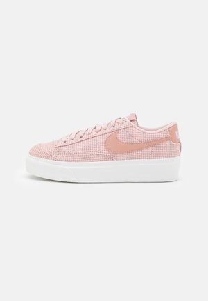 Lyserød low-top sneaker med tekstureret stof, matchende lyserød Nike-swoosh, snørelukning og tyk hvid sål mod en ensfarvet baggrund.