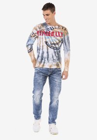 Langärmliges Batik-Shirt in Beige, Blau und Grau mit grafischem Text. Kombiniert mit hellblauen Jeans und weißen Turnschuhen.