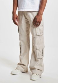 Pantaloni cargo beige con più tasche, vestibilità rilassata e cuciture in vista; indossati con sneakers bianche e una maglietta bianca corta.