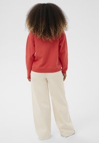 Oseba s kodrasto frizuro, oblečena v rdečo dolgoslečeno majico in ohlapne bež hlače, stoji obrnjena stran od običajne ozadje.