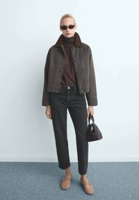 Persona vestida con chaqueta oscura corta, jersey de cuello alto marrón, vaqueros negros de tiro alto, mocasines marrones, gafas de sol, sosteniendo un pequeño bolso marrón.