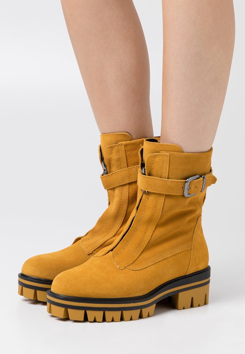 Bottes-cheville en daim jaune avec semelles épaisses crantées, fermeture éclair à l'avant et sangles à boucle latérales portées sur des jambes nues.