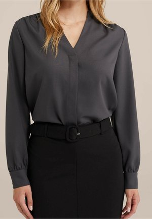 Femme portant un chemisier à manches longues gris foncé rentré dans une jupe noire avec une ceinture noire assortie.