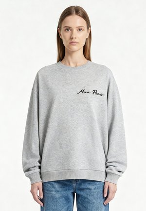 Jeune femme aux cheveux raides portant un sweat-shirt gris clair avec "Mira Paris" brodé en noir sur la poitrine et un jean bleu.