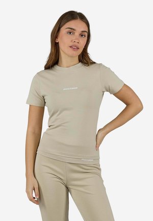 Jeune femme portant une chemise beige à manches courtes et un pantalon assorti, debout avec une main sur la hanche, sur fond blanc uni.