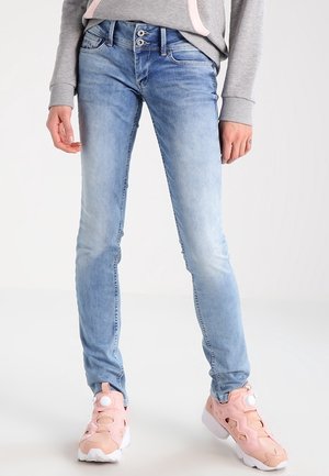 Personne portant un jean skinny bleu clair et des baskets roses avec un sweat-shirt gris, debout sur un fond uni clair.