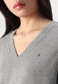 Maglione grigio a scollo a V realizzato in materiale a maglia. Presenta un piccolo logo ricamato in rosso, bianco e blu sulla zona del petto sinistro.