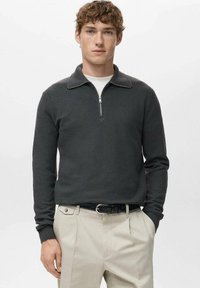 Pull-over à fermeture éclair gris foncé en tissu tricoté texturé avec col, associé à un pantalon beige et une ceinture noire.