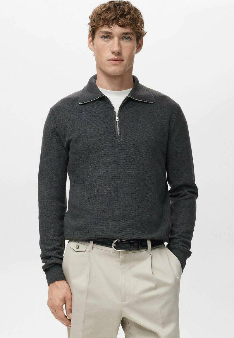 Pull-over à fermeture éclair gris foncé en tissu tricoté texturé avec col, associé à un pantalon beige et une ceinture noire.