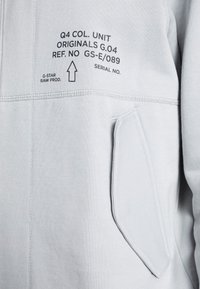 Ljusgrå sweatshirt med en dragkedjeficka, med tryckt text på vänster sida och synliga sömmar längs sömmarna.