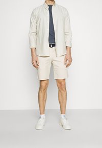 Beige nappatakki, tummansininen t-paita alla, vaaleat shortsit, valkoiset urheilukengät. Suorat pinnat, rento muotoilu, minimalistinen tyyli.