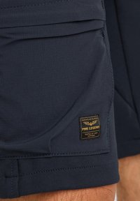 Marineblauwe shorts van een gestructureerde stof, met een opvallend zwart en gouden label aan de zijkant en gedetailleerde stiksels langs de zoom.