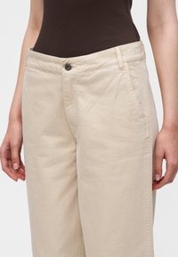 Beige bukser med høy midje, knapp foran og lommer, brukt med en mørk ribbestrikket ermeløs topp, avkortet i midjen.