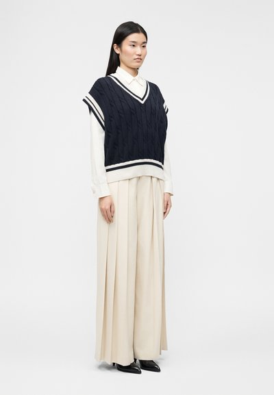 Femme portant un pull sans manches en maille torsadée bleu marine par-dessus une chemise blanche à boutons et un pantalon large beige plissé avec des chaussures noires pointues.