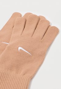Bež pletene rokavice z belim Nike swoosh logotipom na hrbtni strani vsake rokavice, položene ravno na beli površini.