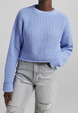 Personne portant un pull en maille épaisse bleu clair et un jean gris clair déchiré, une main dans la poche, sur un fond neutre.