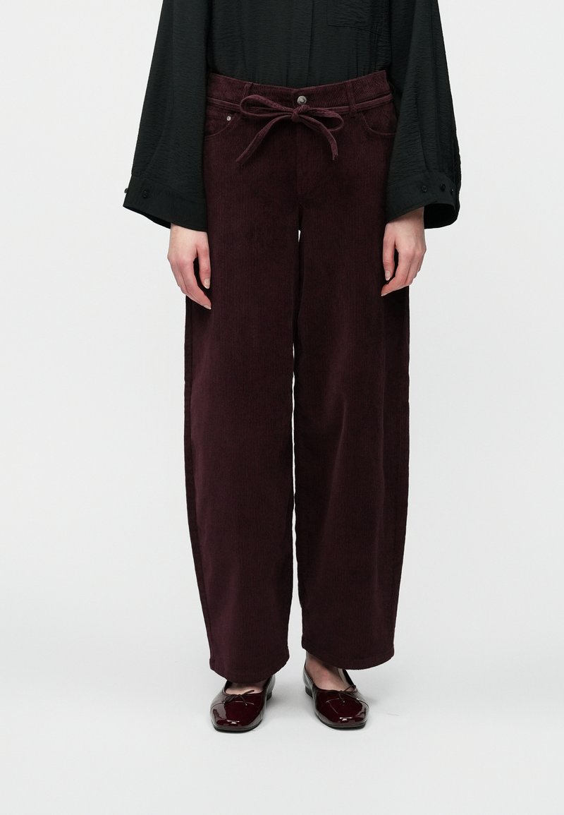 Bordo aksominiai plačių kojų džinsai su juostos raišteliu ir priekinėmis kišenėmis, turintys tekstūrinį paviršių ir subtilius vertikalius raukšlelius.