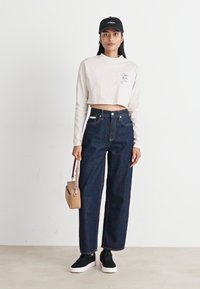 Haut léger beige crop à manches longues, jeans taille haute en denim foncé à jambes larges, baskets noires et une casquette noire au design simple.