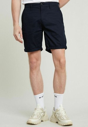 Homme portant un short noir retroussé, des chaussettes blanches et des baskets beiges épaisses, debout devant un fond uni.