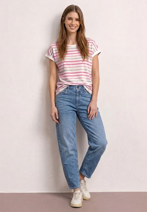 Młoda kobieta uśmiechnięta, ubrana w biało-różowy paskowany t-shirt, jasnoniebieskie dżinsy i białe trampki, stoi na tle jednolitej ściany.