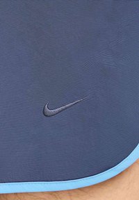 Tamsiai mėlyni sportiniai šortai su šviesiai mėlynu apvadu, turintys išsiuvinėtą juodą Nike swoosh logotipą. Audinys turi tekstūruotą paviršių.