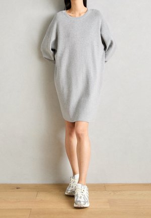 Vrouw draagt een losse, lange mouwen, lichtgrijze trui-jurk en witte sneakers, staand binnen tegen een effen lichte muur.