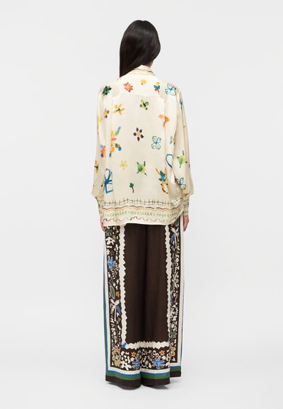 Γυναίκα με μακριά σκούρα μαλλιά φοράει φαρδιά κρεμ μπλούζα με πολύχρωμα αφηρημένα λουλουδένια σχέδια και φαρδιά καφέ παντελόνια με διακοσμητικά περιθώρια.