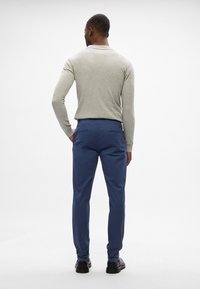 Helle graue Langarmbluse kombiniert mit blauen Hosen. Die Hose hat Gesäßtaschen mit Klappen und ein gerades Bein-Design. Schwarze Schuhe getragen.