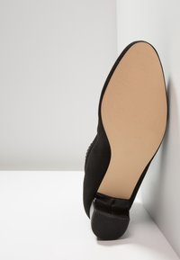 Chaussure en tissu noir avec une tige texturée, un bout arrondi et un talon bas noir brillant. Semelle extérieure en caoutchouc marron avec un motif antidérapant.