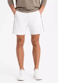 Shorts en coton blancs avec des bandes latérales noires, dotés d'une ceinture élastique et de poches latérales. Portés avec des baskets blanches.