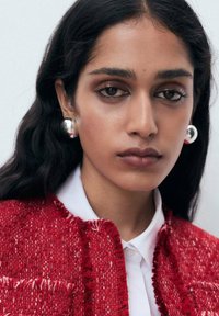 Rote Tweed-Jacke mit ausgefransten Kanten, darunter ein weißes Hemd und große silberne Ohrringe. Glatte Textur, Nahaufnahme-Porträt.
