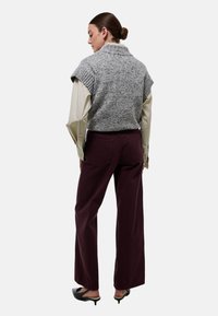 Un pull sans manches en tricot gris avec des accents côtelés, porté sur une chemise à manches longues rayée. Un pantalon large bordeaux et des chaussures noires.