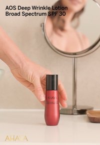 Röd flaska med AHAVA Deep Wrinkle Lotion med svart kork, märkt med Broad Spectrum SPF 30, hålls nära en spegel i en badrumsmiljö.