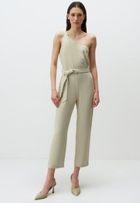 Jumpsuit asimmetrico a spalla unica in tessuto beige chiaro, con cintura in vita, gambe larghe e texture liscia.