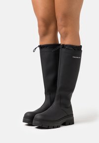 Bens iförda svarta kalvhöga boots från Calvin Klein med tjocka sulor och dragsko stängningar högst upp mot en vit bakgrund.