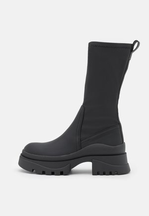 Schwarzer kniehoher Stiefel aus glattem Material, mit einer dicken, profilierten Sohle und einer Schlaufe oben für einfaches Anziehen.