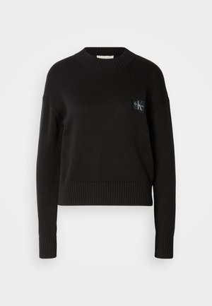 Suéter de punto negro con cuello redondo, puños acanalados y dobladillo. Presenta un pequeño parche de logo cuadrado en el lado izquierdo del pecho. Corte casual.