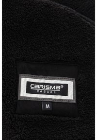 Črna flisna tkanina z etiketo, na kateri je napisano "CARISMA CASUAL" z belimi črkami na sivem ozadju, in oznako velikosti "M." Mehak tekstura in preprost dizajn.