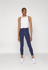 Hvid atletisk crop top med sideskår og sorte detaljer, parret med marineblå højtaljede leggings. Model iført hvide sneakers.