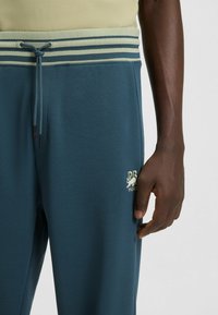 Teal sweatpants with a green striped waistband, featuring a front drawstring, smooth texture, and an embroidered logo near the hem.