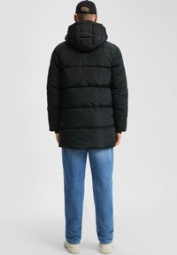 Mann in einer schwarzen Steppjacke mit Kapuze, blauen Jeans, weißen Stiefeln und einer schwarzen Mütze, der sich vor einem schlichten Hintergrund abwendet.