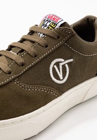 Baskets Vans marron avec panneaux en daim et tissu, logo blanc cousu sur le côté, lacets marron, et semelle en caoutchouc blanche avec motif antidérapant.