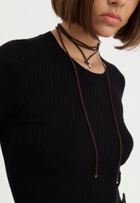 Haut noir côtelé avec un col rond, orné d'un cordon marron avec un accent métallique au cou. Texture lisse.