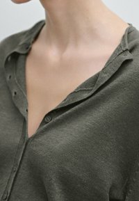 Gebreid shirt in donkergroen, met een knoopsluiting en een V-hals ontwerp. De stof heeft een zachte textuur met een subtiel geribd patroon.