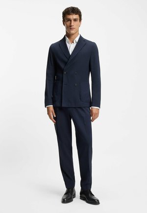 Traje azul marino de doble botonadura con solapa con muesca, botones contrastantes, dos bolsillos delanteros, combinado con camisa blanca y zapatos negros.
