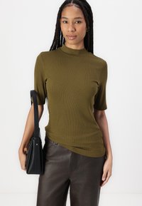 Olivenfarget ribbestrikket topp med kort erm og høy hals, sammen med mørkebrune lærbukser. Modellen holder en svart veske. Enkelt design.